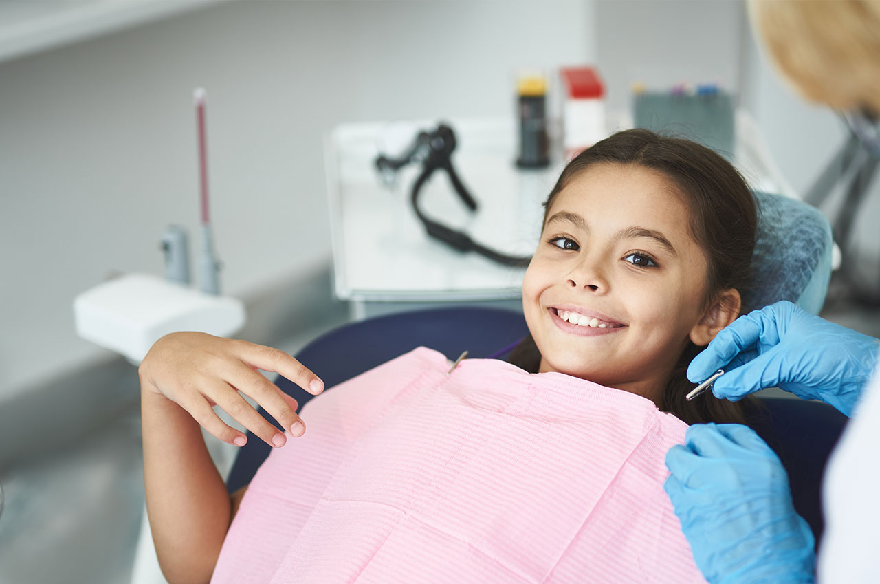A young girl sitting in a dental chair with her mouth open, receiving dental care from a professional wearing blue gloves and a face mask.